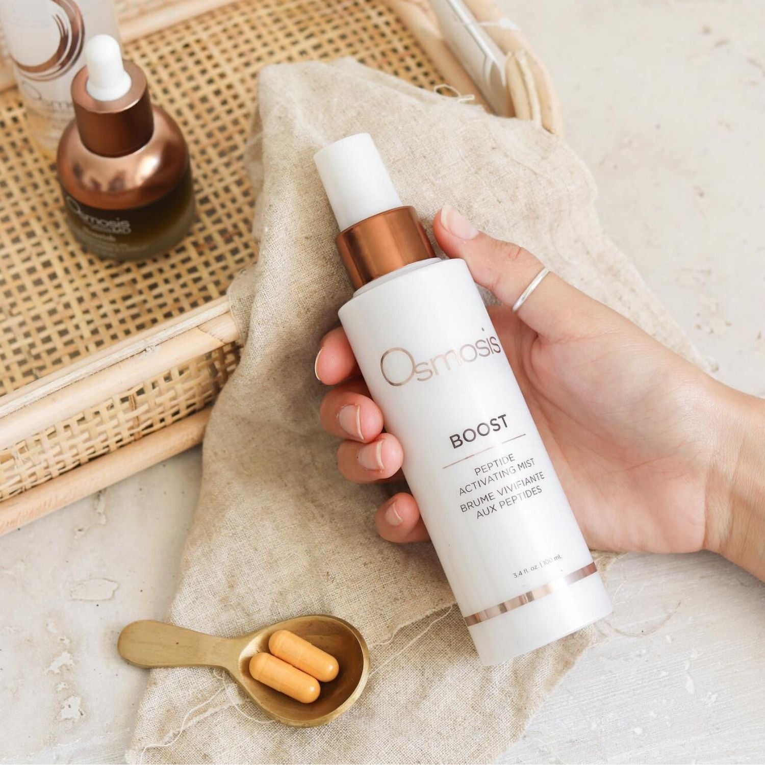 Model hand holding a bottle of boost mist on beige linens with other Osmosis skin care products around