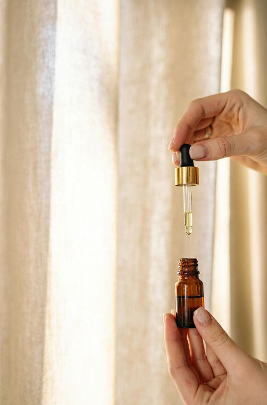Hand holding a dropper bottle over a small brown glass bottle with a beige curtain background