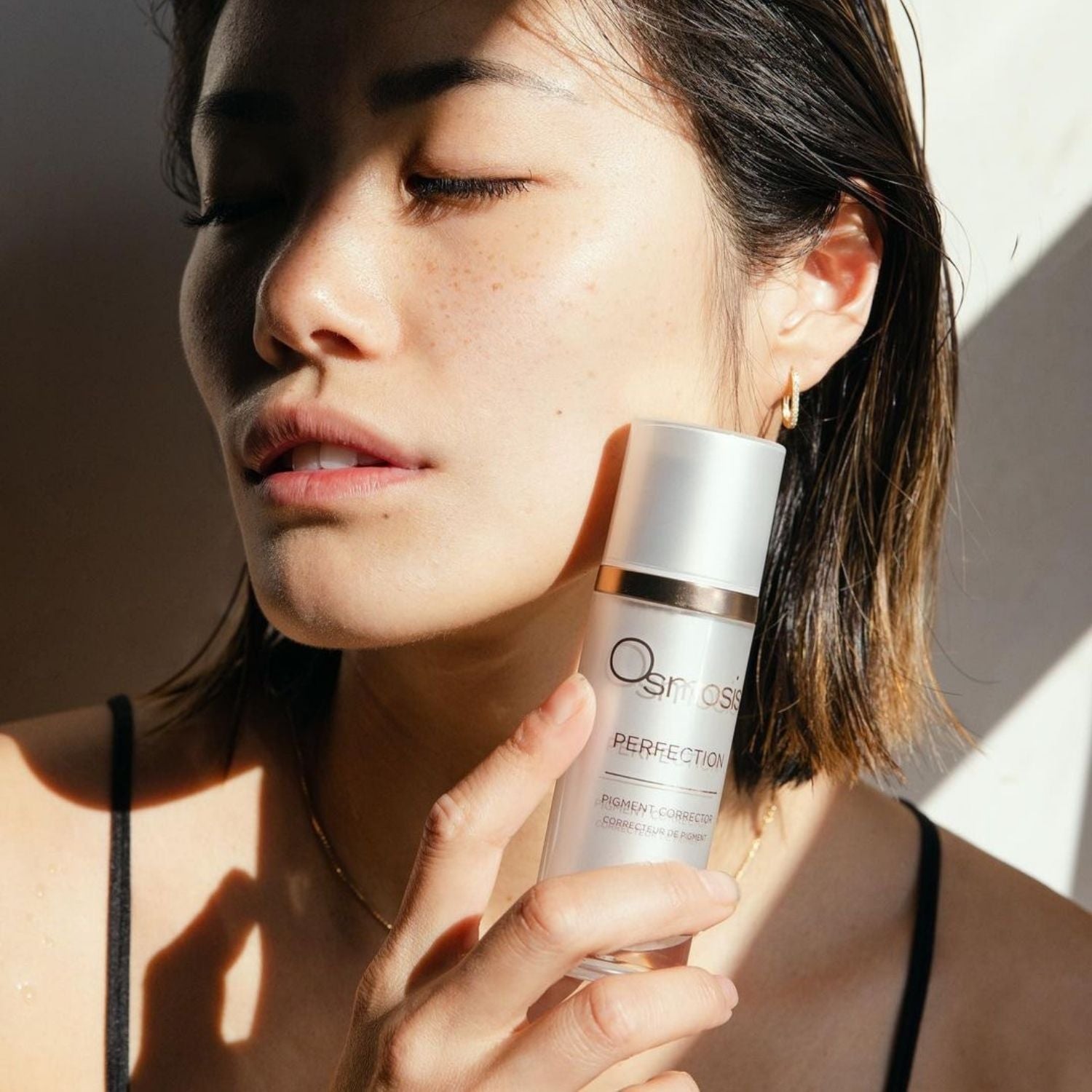 Close up of a model with healthy skin holding a bottle of osmosis perfection pigment corrector near her face