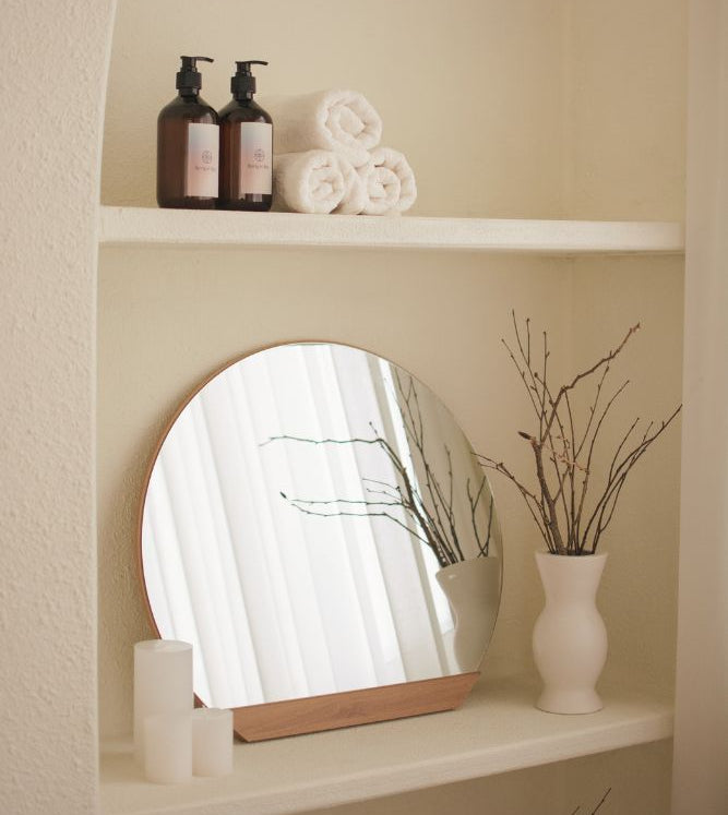 Round mirror on a shelf with bottles and towels in a bathroom setting